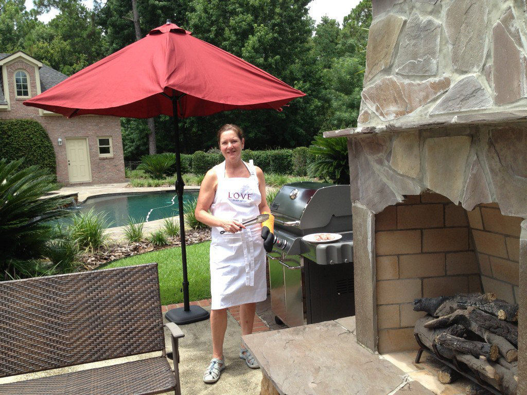 Nikki Landau in a LOVE apron outside her home in Alabama with a large stone fireplace and pool in the background.