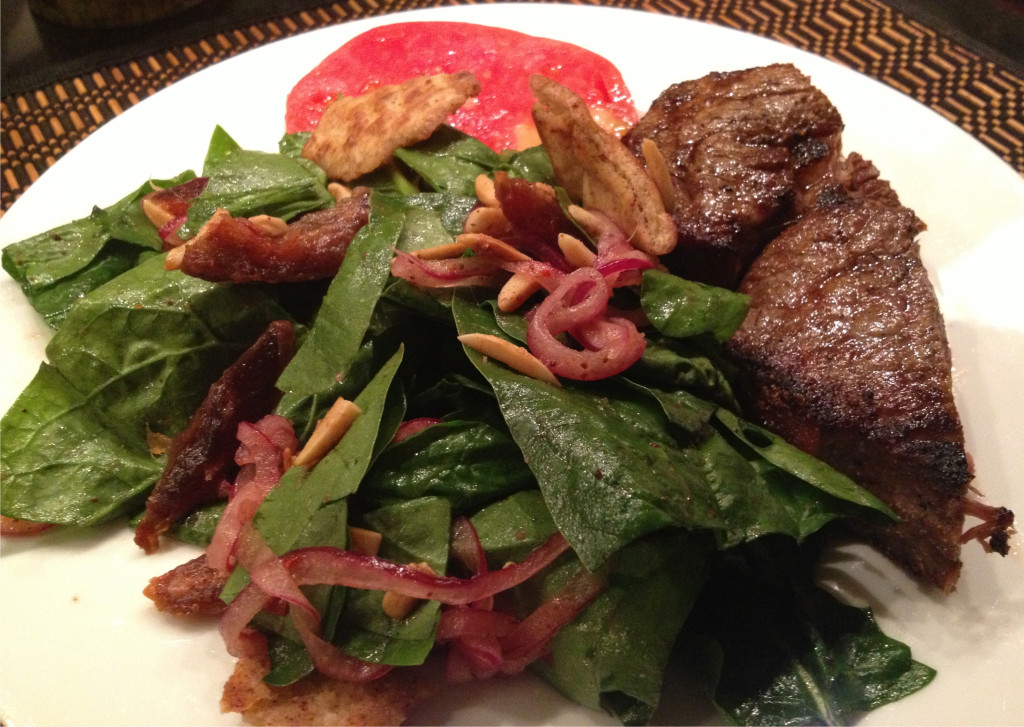 Spinach salad with dates & almonds on a plate with broiled rib steak and an heirloom tomato slice.