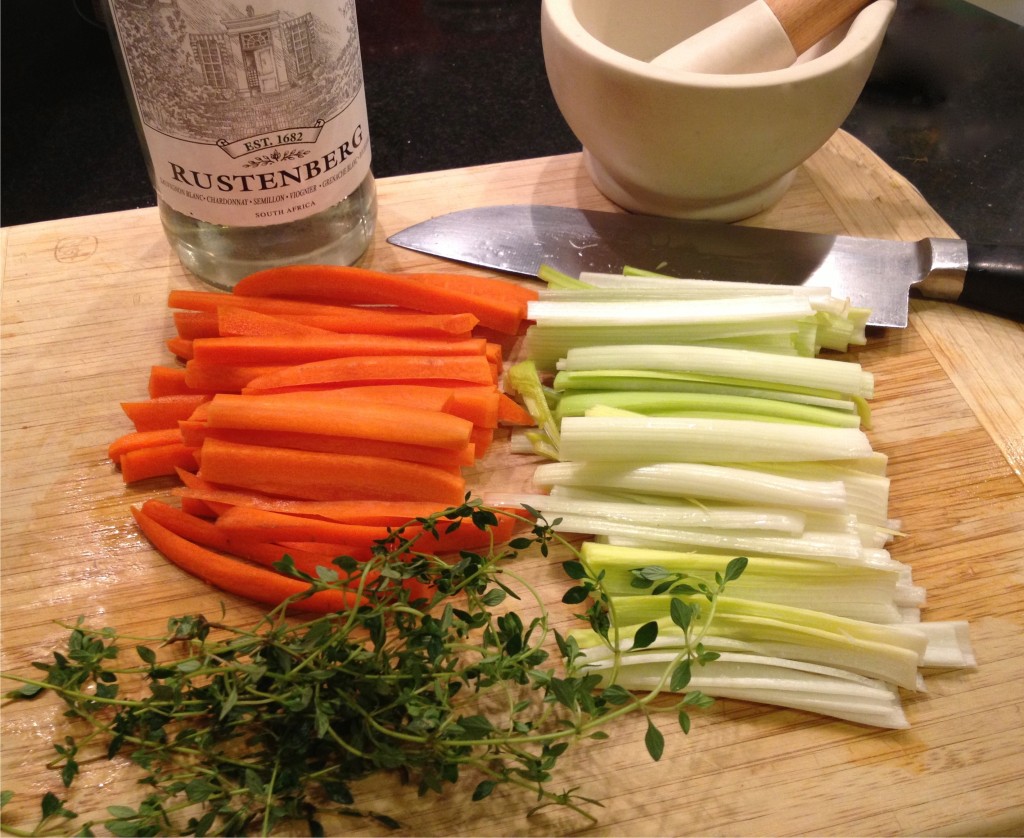 Julienned carrots and leeks with fresh thyme on a cutting board along with a bottle of white wine and mortar and pestle.
