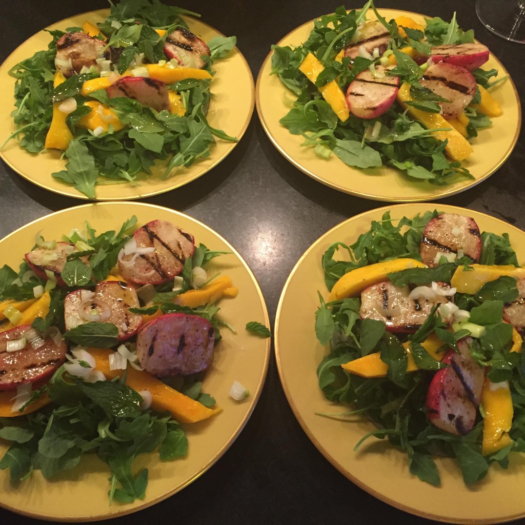 Baby arugula salad with grilled radishes, mango, mint and scallions on four yellow rimmed plates.