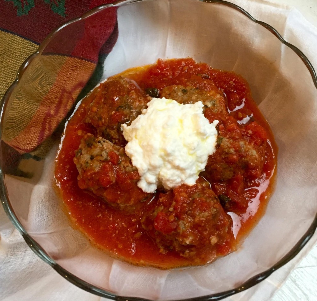 Meatballs made with veal & lamb in a rich tomato sauce topped with ricotta cheese in a glass bowl.
