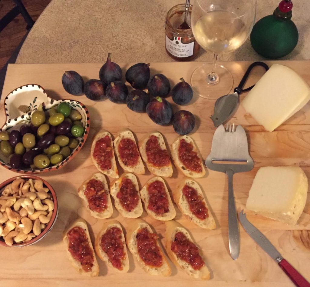 Bruschetta‎, olives, fresh figs, cashews, and 2 kinds of Pecorino on a wooden board with a glass of white wine.