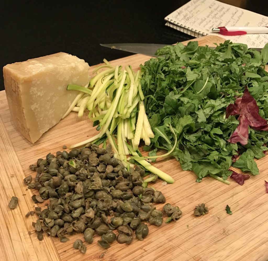 Hunk of Parmigiano cheese, chopped hearty greens, drained and chopped capers, and julienned zucchini on a wooden board, handwritten recipe in background.