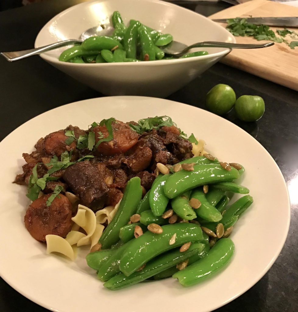 Lamb stew over egg noodles with blanched sugar snap peas topped with pepitos in a white bowl.