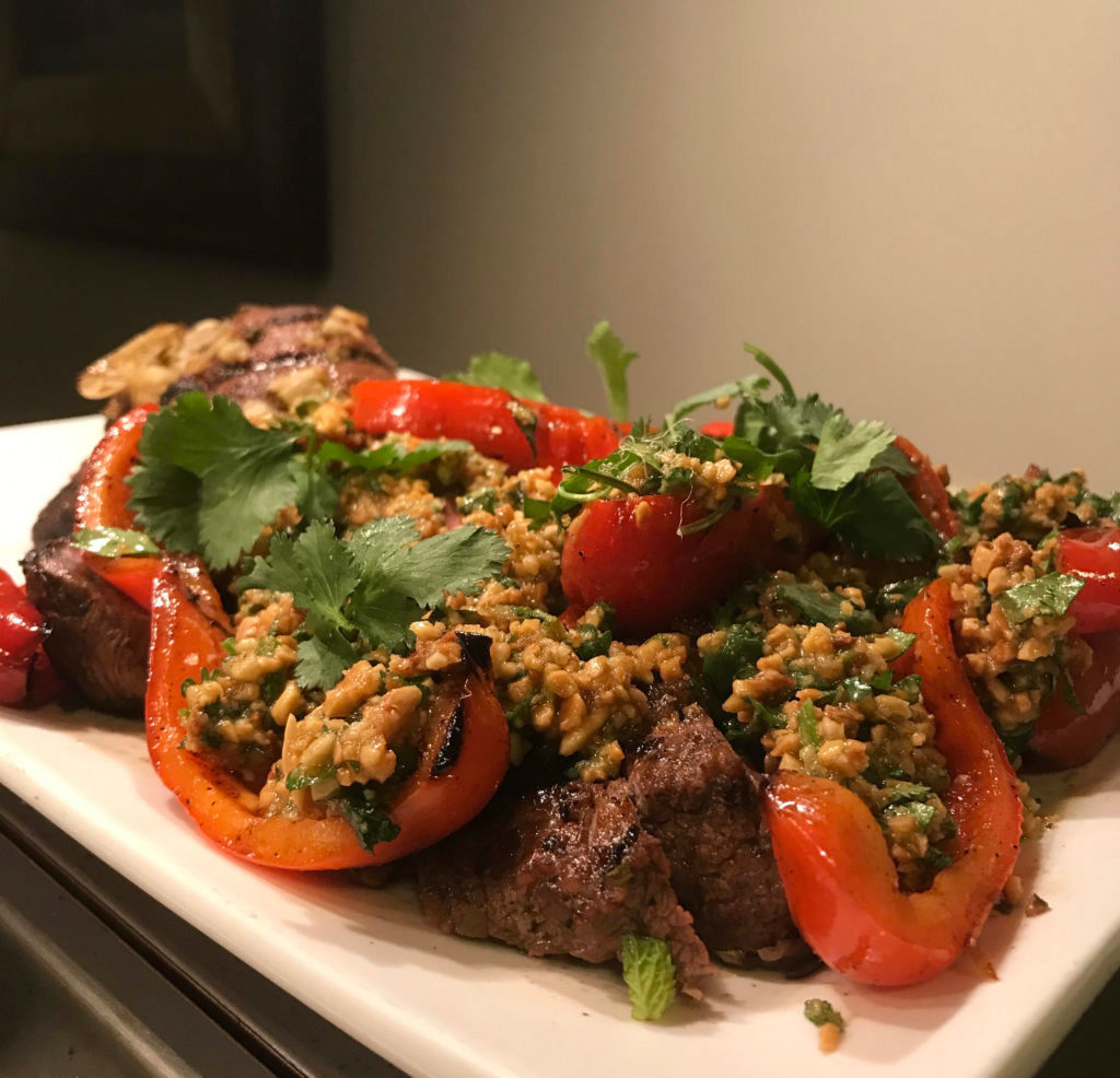Ottolenghi Grilled Leg of Lamb recipe with red peppers and almond sauce on a white platter, garnished with cilantro.